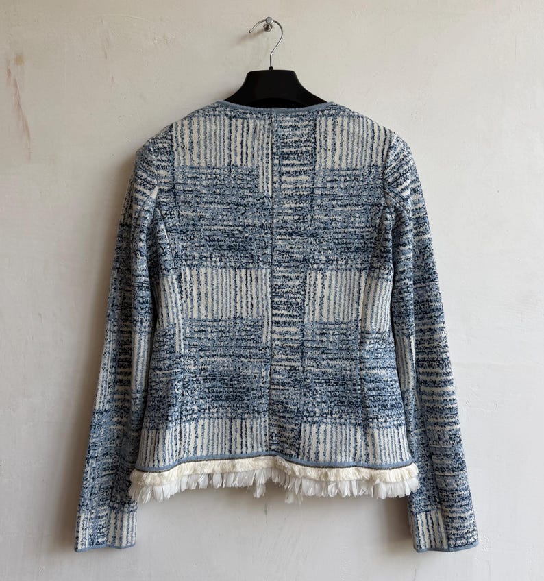 May include: A long-sleeved jacket featuring a blue and white textured plaid pattern. The garment has a round neckline, a straight hemline, and is finished with a decorative fringe trim along the bottom edge. The jacket is displayed on a hanger.