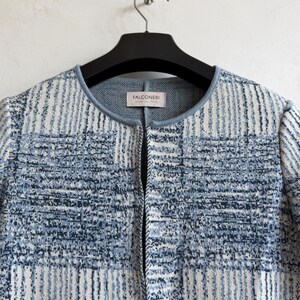 May include: A long-sleeved knit jacket featuring a blue and white checkered pattern with a textured finish. The garment has a round neckline with a solid light blue trim and a front opening. A label inside the neck reads Falconeri made in Italy.