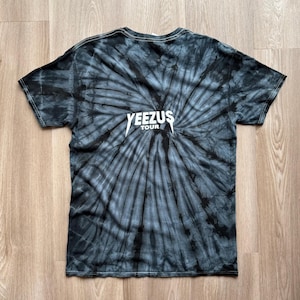 Może przedstawiać: Czarna i szara koszulka typu tie-dye z białym napisem "YEEZUS TOUR". Koszulka ma spiralny wz&oacute;r promieniujący ze środka. Koszulka z kr&oacute;tkim rękawem jest wykonana z miękkiego materiału.