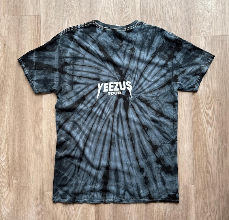 Może przedstawiać: Czarna i szara koszulka typu tie-dye z białym napisem "YEEZUS TOUR". Koszulka ma spiralny wz&oacute;r promieniujący ze środka. Koszulka z kr&oacute;tkim rękawem jest wykonana z miękkiego materiału.