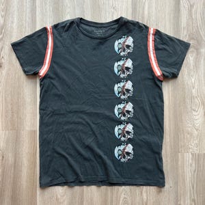 May include: A dark gray t-shirt with a vertical row of skull graphics on the right side. The shirt has red and white stripes on the sleeves. The text "ALLSAINTS" is printed on the shirt.