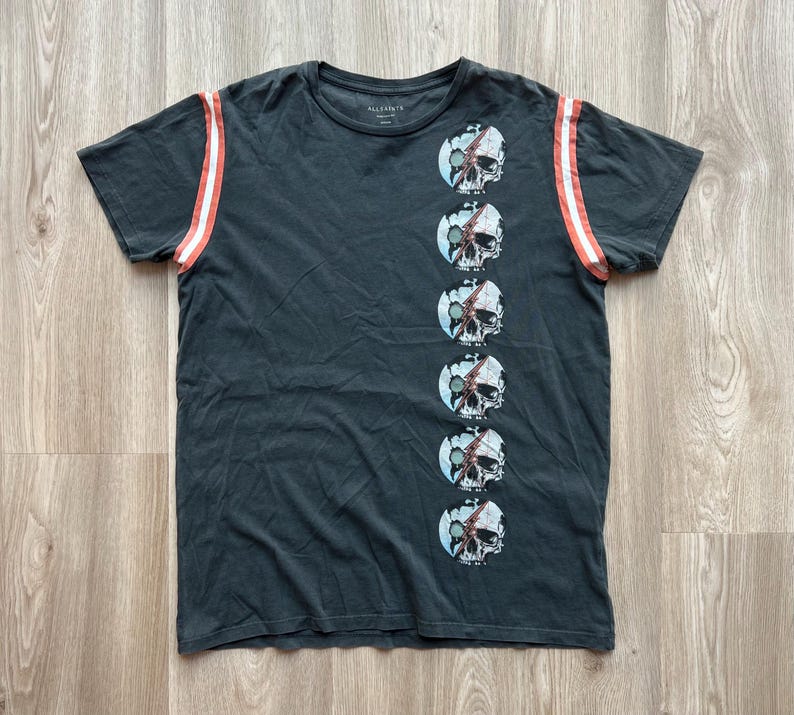 May include: A dark gray t-shirt with a vertical row of skull graphics on the right side. The shirt has red and white stripes on the sleeves. The text "ALLSAINTS" is printed on the shirt.