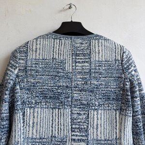 May include: A long-sleeved jacket featuring a textured, woven pattern in shades of blue, white, and dark navy. The garment has a round neckline with a solid blue trim and a vertical seam running down the center back, displayed on a black hanger.