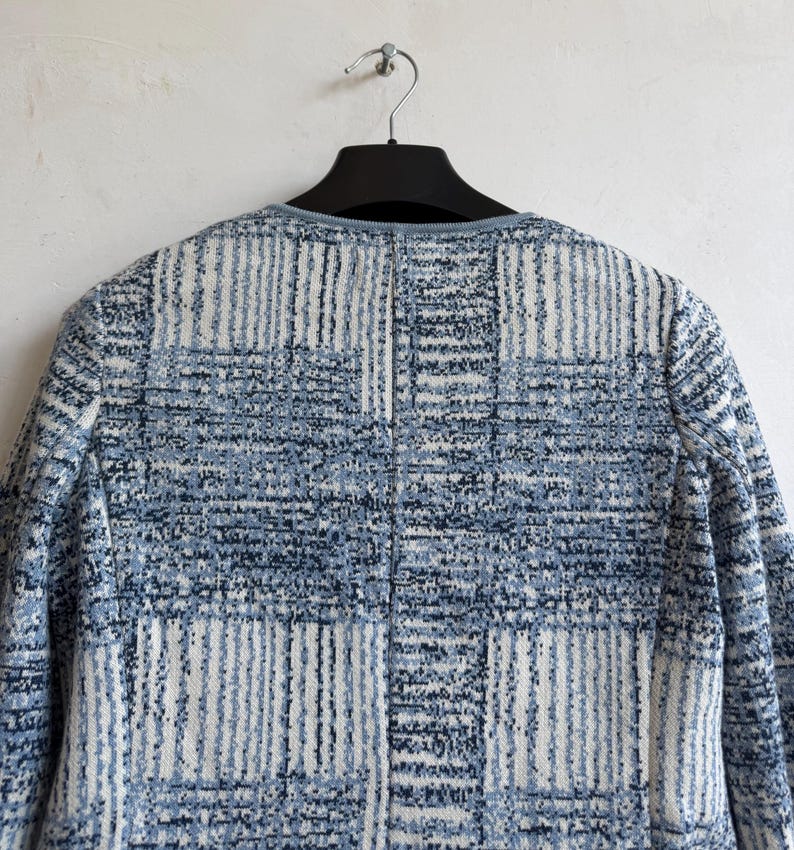 May include: A long-sleeved jacket featuring a textured, woven pattern in shades of blue, white, and dark navy. The garment has a round neckline with a solid blue trim and a vertical seam running down the center back, displayed on a black hanger.