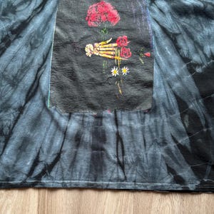 Może przedstawiać: Czarna i szara koszulka typu tie-dye z grafiką przedstawiającą szkielet dłoni trzymającą czerwone r&oacute;że i stokrotki. Grafika znajduje się na czarnej prostokątnej naszywce. Koszulka wykonana jest z miękkiego materiału.