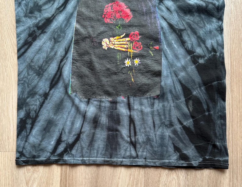 Może przedstawiać: Czarna i szara koszulka typu tie-dye z grafiką przedstawiającą szkielet dłoni trzymającą czerwone r&oacute;że i stokrotki. Grafika znajduje się na czarnej prostokątnej naszywce. Koszulka wykonana jest z miękkiego materiału.