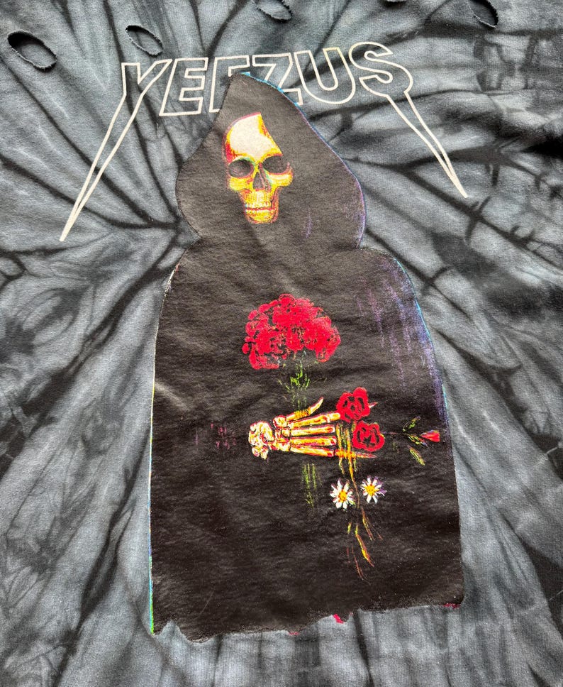 Może przedstawiać: Koszulka typu tie-dye z napisem "YEEZUS" w kolorze białym u g&oacute;ry. Koszulka przedstawia grafikę ponurego żniwiarza trzymającego czerwone r&oacute;że i stokrotki. Żniwiarz ma głowę czaszki i nosi czarny płaszcz.