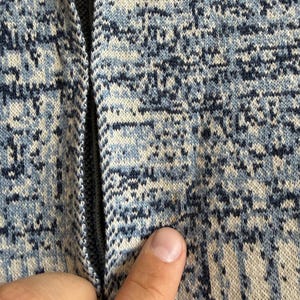 May include: A knit fabric garment featuring a mottled pattern of dark blue, light blue, and off-white threads. The material has a textured appearance with a vertical seam running down the center, displaying a small, faint orange spot on the surface.