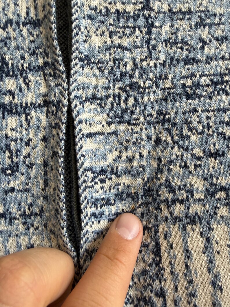 May include: A knit fabric garment featuring a mottled pattern of dark blue, light blue, and off-white threads. The material has a textured appearance with a vertical seam running down the center, displaying a small, faint orange spot on the surface.