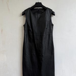 May include: A sleeveless black dress made of a smooth material, featuring a round neckline and a visible vertical zipper closure at the upper back. The garment has horizontal seam detailing across the chest and mid-section, creating a structured silhouette.