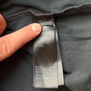 May include: Black care tag with white text. The tag includes size information, "M 185/100A", and care instructions in French and English. The tag is attached to a black garment.