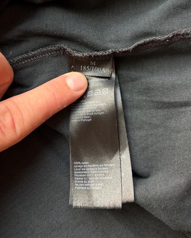 May include: Black care tag with white text. The tag includes size information, "M 185/100A", and care instructions in French and English. The tag is attached to a black garment.