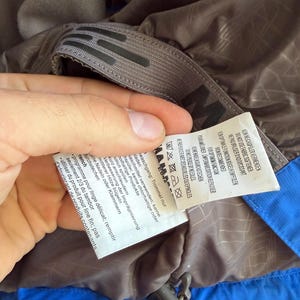 May include: A blue and brown jacket interior featuring a grey hanging loop with a black logo and a white care instruction label. The label includes text in multiple languages, laundry symbols, and instructions regarding fabric softener and ironing.