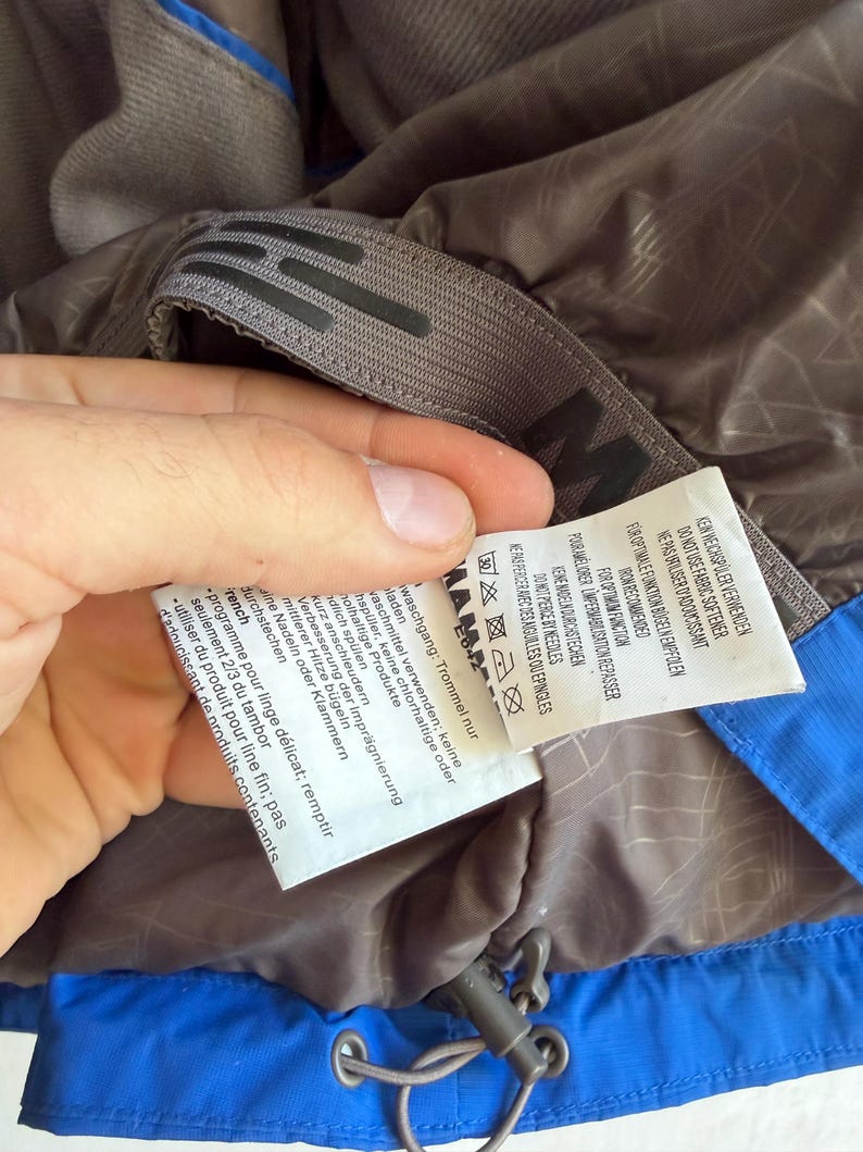 May include: A blue and brown jacket interior featuring a grey hanging loop with a black logo and a white care instruction label. The label includes text in multiple languages, laundry symbols, and instructions regarding fabric softener and ironing.
