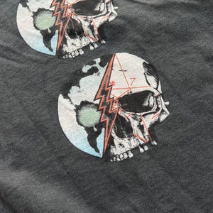 May include: Gray t-shirt with a graphic print of a skull with a lightning bolt and geometric patterns on a blue and green globe.