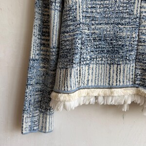 May include: A long-sleeved knit sweater featuring a blue and white plaid pattern with a textured, mottled appearance. The hem is finished with a decorative trim consisting of a thin metallic chain and a layer of white fringe.