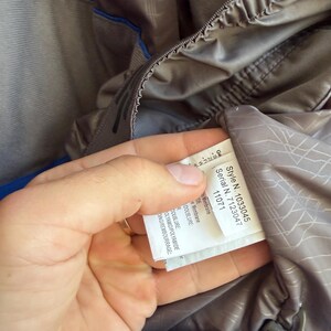 May include: A blue and grey jacket with an interior label displaying Style N. 1033045 and Serial N. 7123047. The garment features a grey lining with a geometric line pattern, a blue outer shell, and a drawstring cord with a plastic toggle fastener.