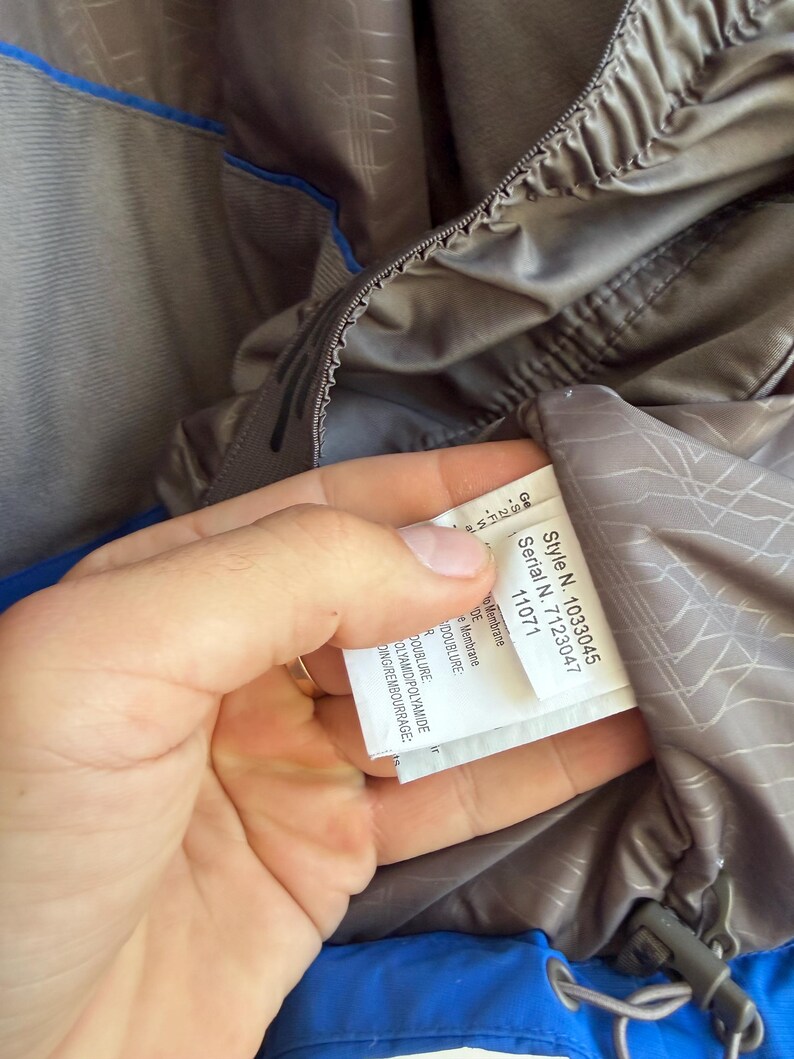 May include: A blue and grey jacket with an interior label displaying Style N. 1033045 and Serial N. 7123047. The garment features a grey lining with a geometric line pattern, a blue outer shell, and a drawstring cord with a plastic toggle fastener.