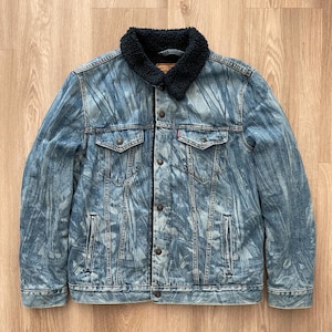 Può includere: Una giacca di jeans blu con fodera in sherpa nera e un motivo astratto sbiadito. La giacca ha una chiusura con bottoni, due tasche sul petto e due tasche anteriori.