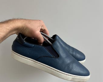 May include: A pair of dark blue leather slip-on shoes with white soles. The shoes have a textured leather upper and a simple design.