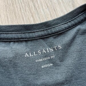 May include: A gray t-shirt with a white tag that says "ALLSAINTS" in a bold font. The tag also says "OVERSIZED FIT" and "MEDIUM".