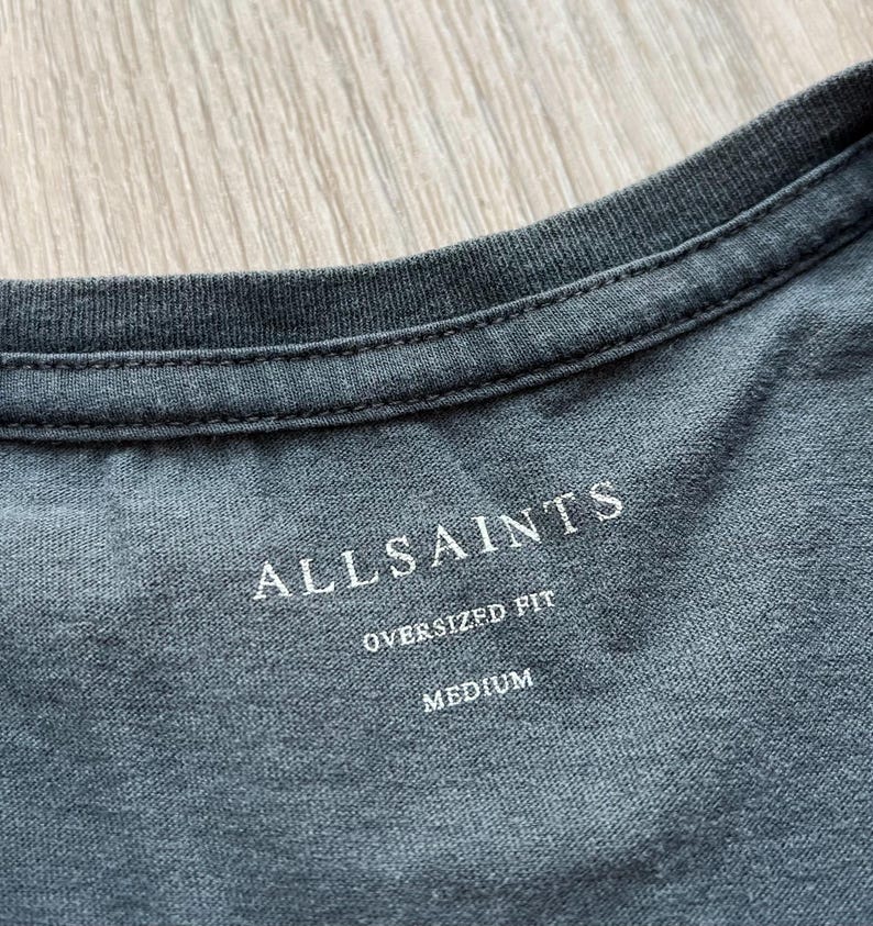 May include: A gray t-shirt with a white tag that says "ALLSAINTS" in a bold font. The tag also says "OVERSIZED FIT" and "MEDIUM".