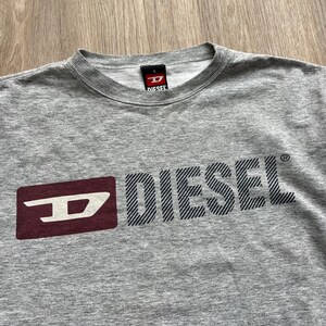 May include: Grey long-sleeved t-shirt with a red and white Diesel logo. The logo is a stylised "D" with the word "Diesel" in black letters with a striped pattern.