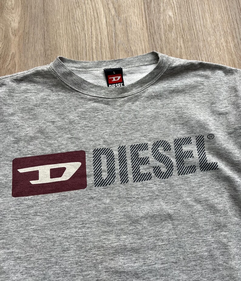 May include: Grey long-sleeved t-shirt with a red and white Diesel logo. The logo is a stylised "D" with the word "Diesel" in black letters with a striped pattern.