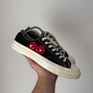 Peut inclure: Baskets basses noires avec une semelle et un embout en caoutchouc blancs. Un cœur rouge avec deux yeux blancs est sur le côté. Les baskets ont des lacets blancs et sont tenues par une main.