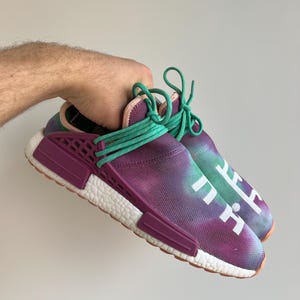 Puede incluir: Zapatillas de deporte de color morado y verde con efecto tie-dye con el texto "HUMAN RACE" en blanco en el lateral. Los cordones son verdes.