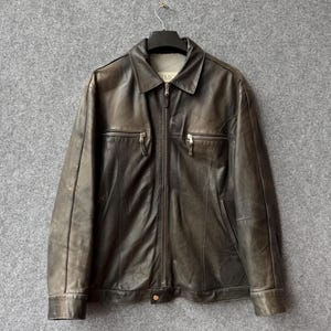 May include: A dark brown leather jacket featuring a full-length front zipper, a standard collar, and two horizontal zippered chest pockets. The jacket has long sleeves with buttoned cuffs and a smooth, slightly distressed finish throughout the leather.