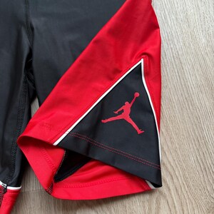 May include: Black and red athletic shorts with white trim. The shorts have a red panel with a silhouette of a person playing basketball. The shorts are made of a stretchy material.