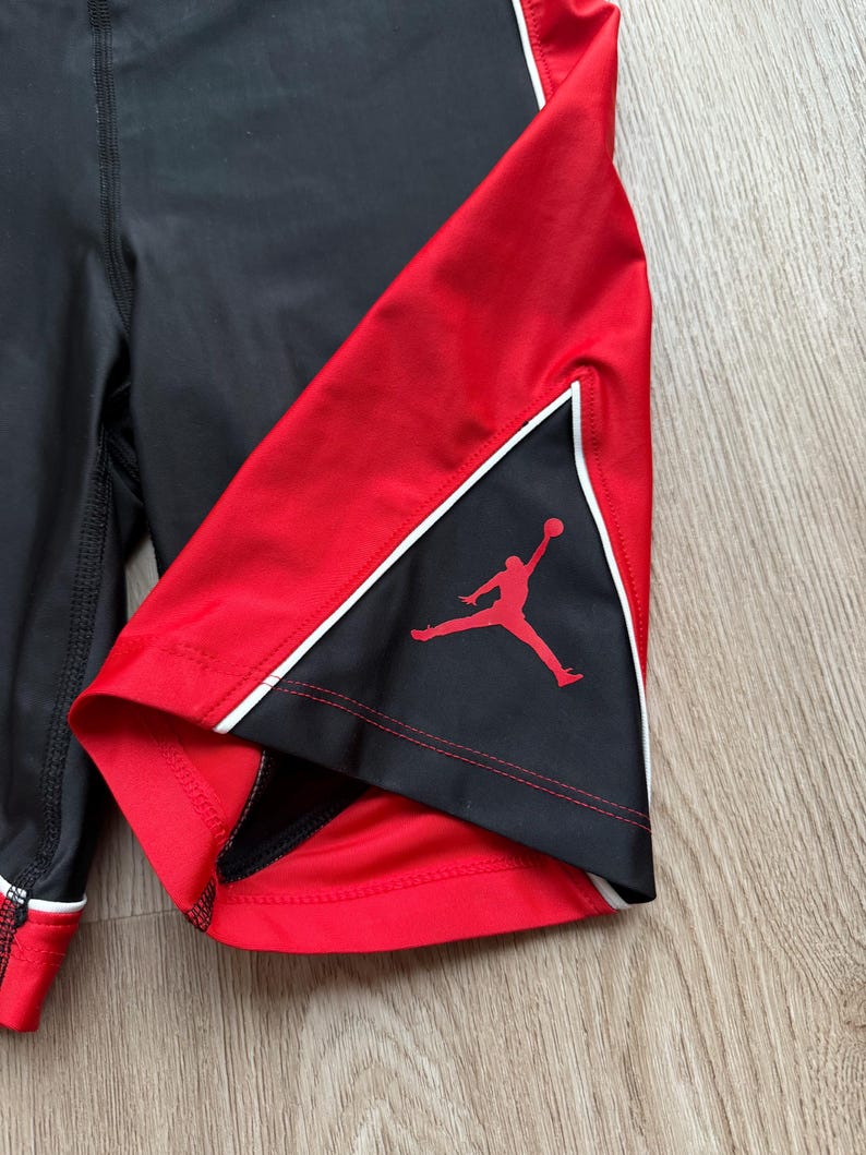 May include: Black and red athletic shorts with white trim. The shorts have a red panel with a silhouette of a person playing basketball. The shorts are made of a stretchy material.
