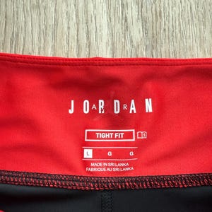 May include: Close-up of red and black athletic leggings. The leggings have a red waistband with the word "JORDAN" in white block letters. The label reads "TIGHT FIT" and "MADE IN SRI LANKA".