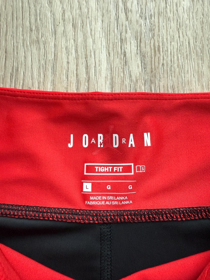 May include: Close-up of red and black athletic leggings. The leggings have a red waistband with the word "JORDAN" in white block letters. The label reads "TIGHT FIT" and "MADE IN SRI LANKA".