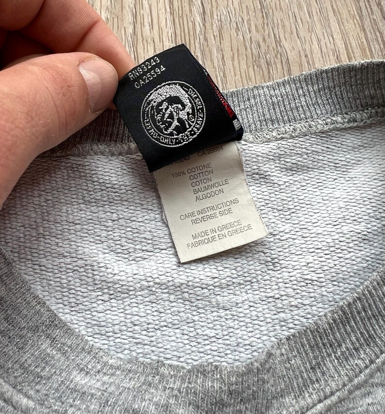 May include: A grey cotton sweatshirt with a Diesel logo tag. The tag features the text "Diesel Only The Brave" and the numbers "RN93243 CA25594". The tag also includes care instructions and information about the fabric composition: 100% cotone, cotton, coton, baumwolle, algodon. The tag also states that the sweatshirt is made in Greece.
