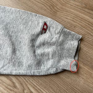 May include: Grey sweatshirt with a red and white logo on the sleeve. The sleeve has a ribbed cuff.