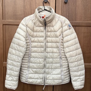 Puede incluir: Chaqueta acolchada de color beige claro con cuello alto y cremallera completa. La chaqueta tiene un diseño acolchado y un pequeño parche con el logotipo en la manga superior izquierda. Colgada de una percha negra.