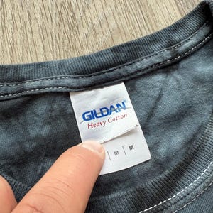 Może przedstawiać: Zbliżenie szarej koszulki tie-dye z białą metką. Na metce widnieje napis "GILDAN Heavy Cotton" w niebieskim i czerwonym kolorze. Koszulka jest w rozmiarze M.