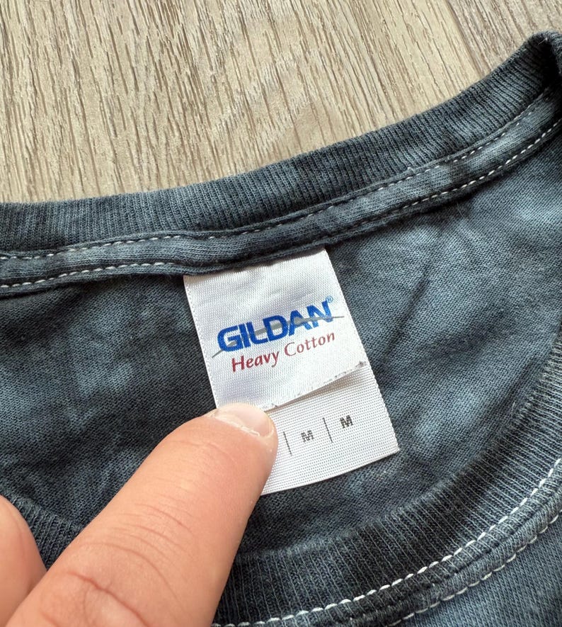 Może przedstawiać: Zbliżenie szarej koszulki tie-dye z białą metką. Na metce widnieje napis "GILDAN Heavy Cotton" w niebieskim i czerwonym kolorze. Koszulka jest w rozmiarze M.