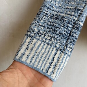 May include: A long-sleeved knit garment sleeve featuring a textured pattern in shades of light blue, dark blue, and white. The cuff displays a distinct vertical striped design that transitions into a mottled, speckled pattern along the arm.