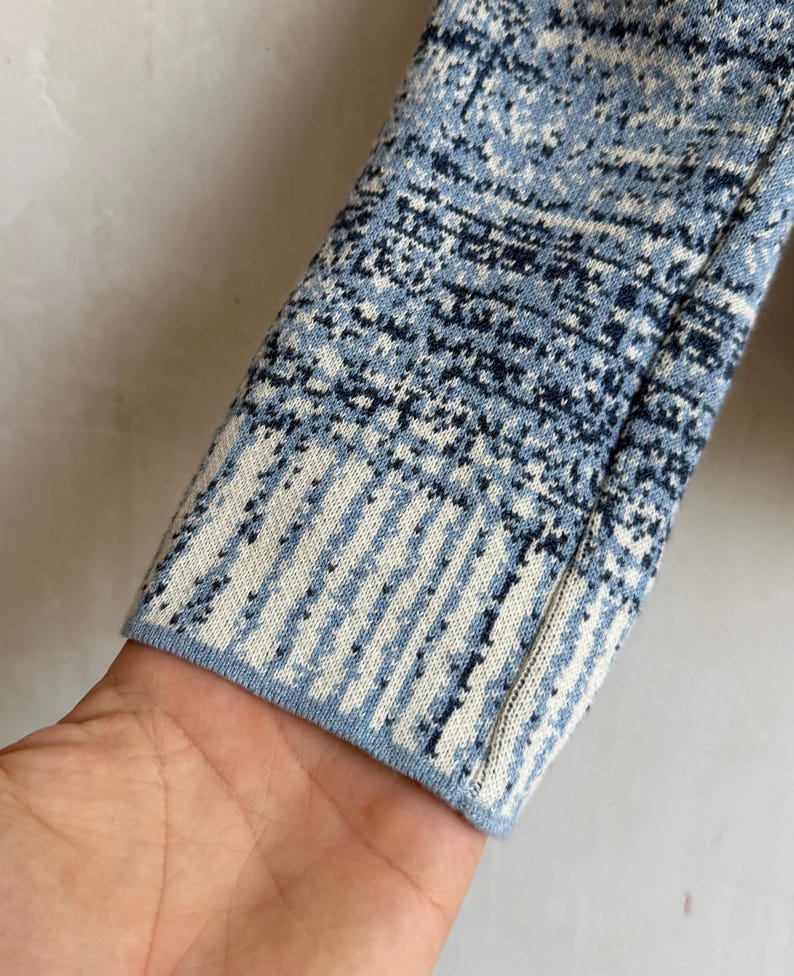 May include: A long-sleeved knit garment sleeve featuring a textured pattern in shades of light blue, dark blue, and white. The cuff displays a distinct vertical striped design that transitions into a mottled, speckled pattern along the arm.