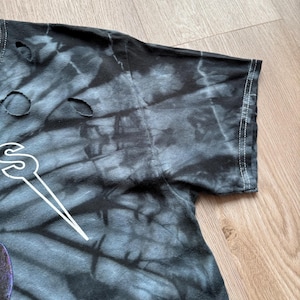 Może przedstawiać: Zbliżenie czarnej i szarej koszulki tie-dye z dziurami. Koszulka ma biały wz&oacute;r graficzny. Widoczny jest rękaw z białymi szwami na krawędzi. Koszulka leży na drewnianej powierzchni.