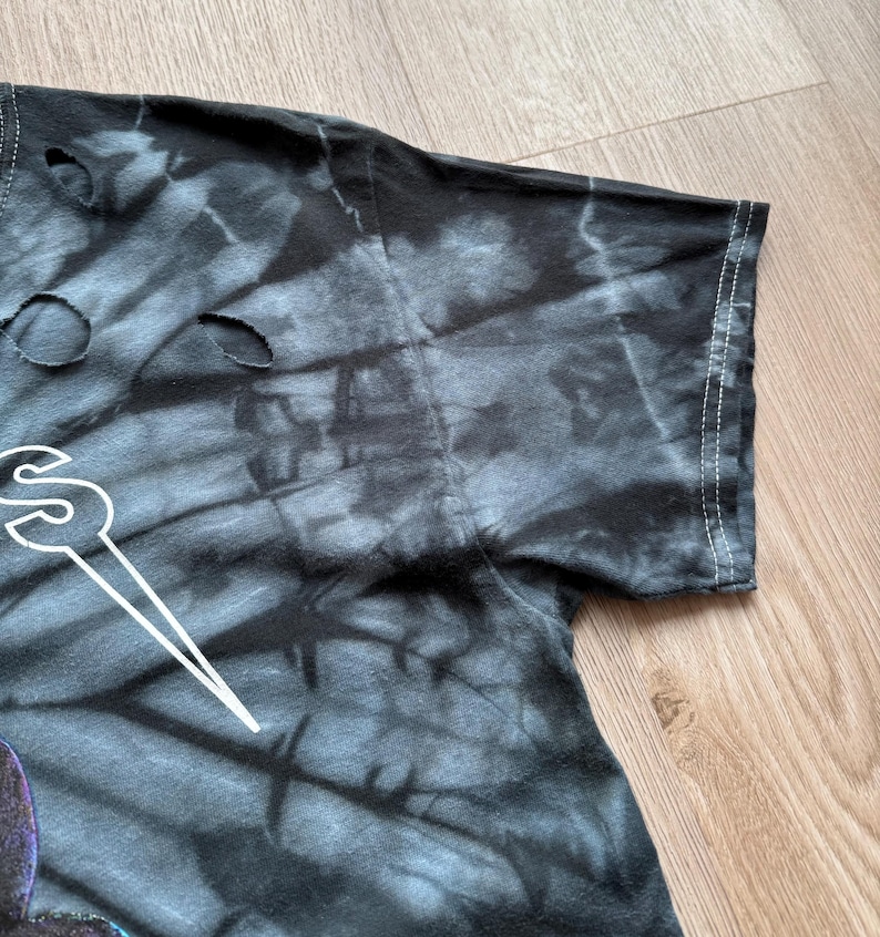 Może przedstawiać: Zbliżenie czarnej i szarej koszulki tie-dye z dziurami. Koszulka ma biały wz&oacute;r graficzny. Widoczny jest rękaw z białymi szwami na krawędzi. Koszulka leży na drewnianej powierzchni.