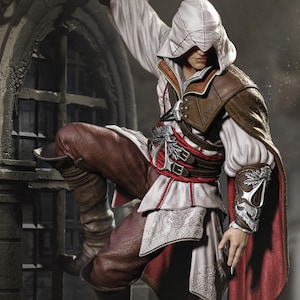 May include: A detailed action figure of a character from the Assassin's Creed video game series. The figure is posed mid-climb, wearing a white hooded cowl, brown leather armor, and red accents. The figure is set against a blurred cityscape background.