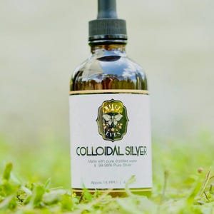 May include: A brown glass bottle with a black dropper top, labeled "COLLOIDAL SILVER." The label features a butterfly design and the text "WILDE EVES." The bottle is set against a blurred green background of grass.