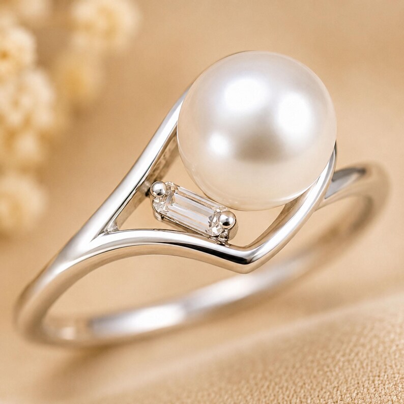 May include: A silver-toned ring featuring a single round white pearl set within a V-shaped band. A small, clear, rectangular baguette-cut gemstone is positioned on the band directly beneath the pearl, adding a subtle accent to the design.