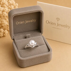 May include: A silver ring featuring a central white pearl flanked by two small baguette-cut stones, presented in a taupe velvet jewelry box with the text Orien Jewelry printed in gold on the interior lid and on an accompanying card.