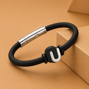 May include: A black silicone bracelet with a silver clasp and a silver "U" charm. The bracelet is oval-shaped and features a magnetic clasp. The charm is a stylized letter "U".