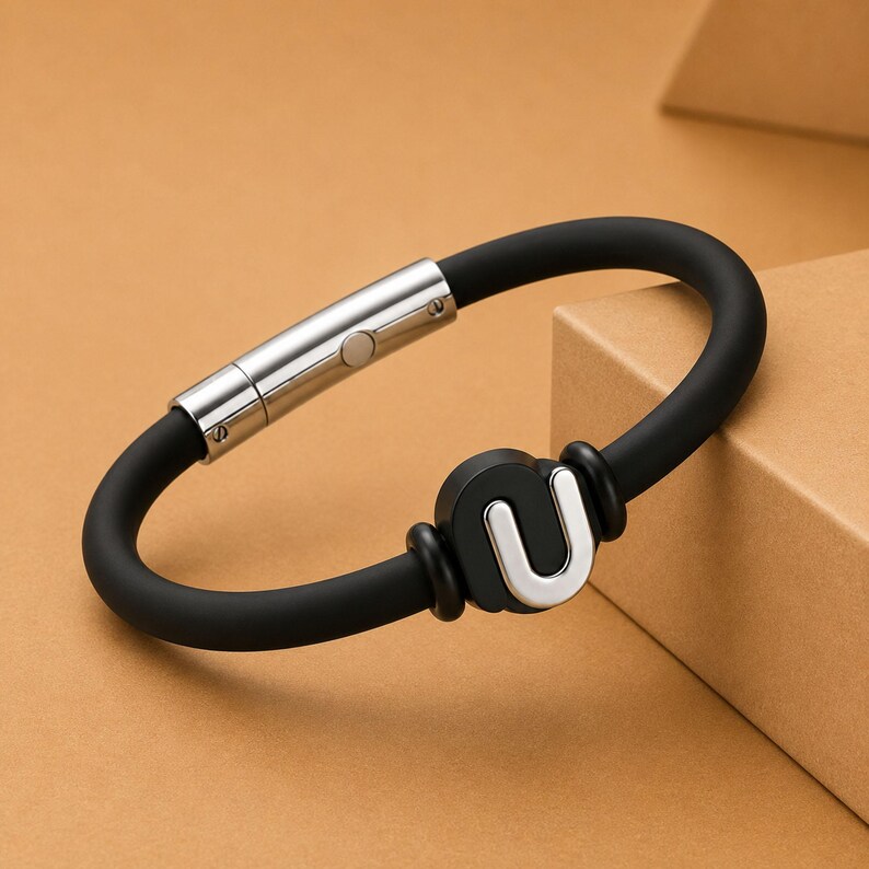 May include: A black silicone bracelet with a silver clasp and a silver "U" charm. The bracelet is oval-shaped and features a magnetic clasp. The charm is a stylized letter "U".